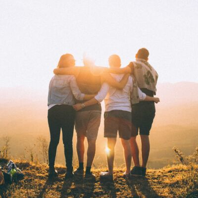 A group of four friends hug each other on a cliff as they watch a sunset.